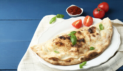 Tasty calzone with basil, tomatoes and sauce on blue wooden table, above view