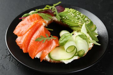 Delicious bagel with salmon, cream cheese, cucumber and avocado on black table, closeup