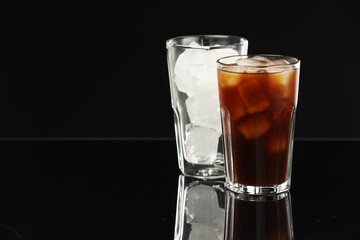 Refreshing iced coffee and ice cubes in glasses on black mirror surface. Space for text