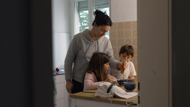Candid domestic lifestyle scene of mother standing in kitchen with her two children preparing meal together. Small brother and sister interacting with parent while she pours honey into bowl