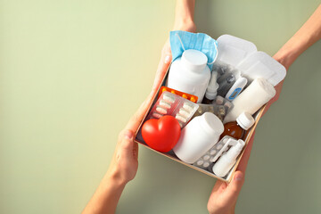 Hands holding a box filled with various medical supplies, including pills, bottles, a heart, a...