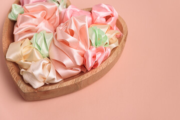 A heart-shaped wooden bowl filled with pastel-colored satin ribbons, curled into delicate roses. The soft pink background creates a romantic and dreamy atmosphere.