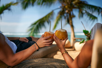 Couple relaxing on beach lounge chairs, enjoying fresh coconut water. Romantic tropical getaway, pair sips natural drinks under palm tree. Ocean view, sunny day, luxury resort, leisure lifestyle.