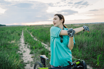 A woman wearing a blue shirt and gloves stretches her arm while standing next to her bicycle. The...