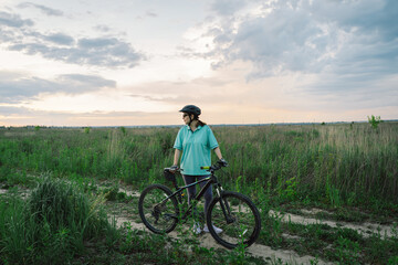 Fototapeta premium A woman wearing a helmet rides a black mountain bike along a dirt path. The path runs through an open grassy field, with houses and trees visible in the background.