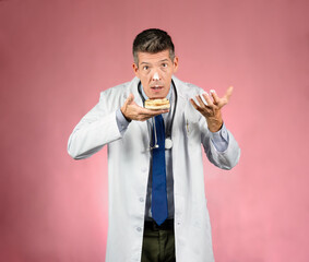 Young handsome doctor with surprised face in gown, holding a cake and a hand with cream and a spot of cream on his nose