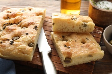 Pieces of delicious focaccia bread with olives, thyme, salt and knife on wooden table, closeup