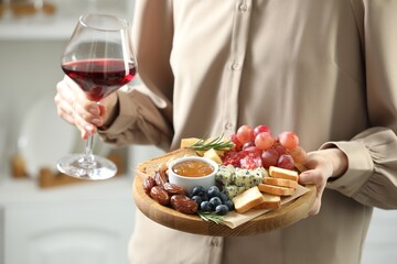 Woman holding board with different types of delicious cheese, other snacks and wine indoors, closeup