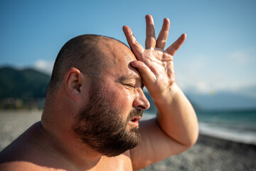 Overweight man suffers heatstroke on beach, shields head with hand. Struggles with heat, wipes...