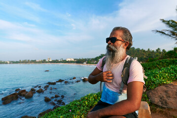 Elderly man, backpack, sits alone on bench gazing at ocean. Reflects on life journey, seeks peace. Senior traveler finds healing, solitude by sea. Nature contemplation enhances mental health.