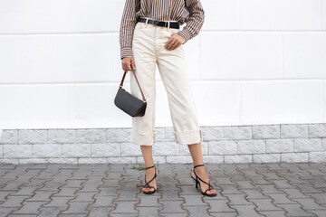 Woman wearing striped shirt, white jeans, black bag, heeled sandals standing outdoor on city...