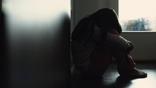 Silhouette of sad depressed child covering face in shame, struggling with mental illness in childhood. Little girl seated at home corridor in gloomy moody scene