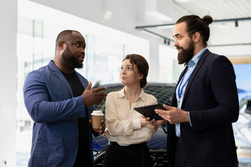 Car sales executive showcasing dealership vehicles to client using tablet, explaining characteristics. Showroom salesperson helping customer choose automobile using digital device, comparing models