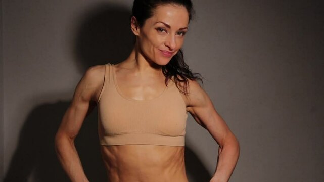 Healthy smiling woman posing and showing abdoman muscule abs, fit curves, attractive body. Slim caucasian female model posing in beige bra in studio low key light on wall background. Sporty lifestyle