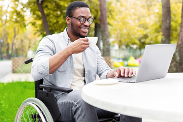 Remote job for impaired people. Cheerful black guy in wheelchair working online, using laptop at...