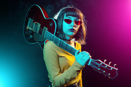 Trendy young hipster woman with curly hair with red guitar in neon lights. Rock musician posing with electrical guitar.