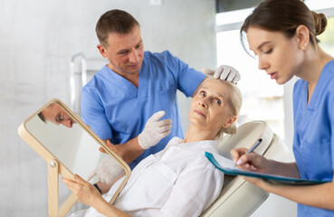 Fototapeta premium Interested elderly woman sitting on examination couch and looking at mirror while professional cosmetologist with female assistant planning and explaining future rejuvenating facial procedure