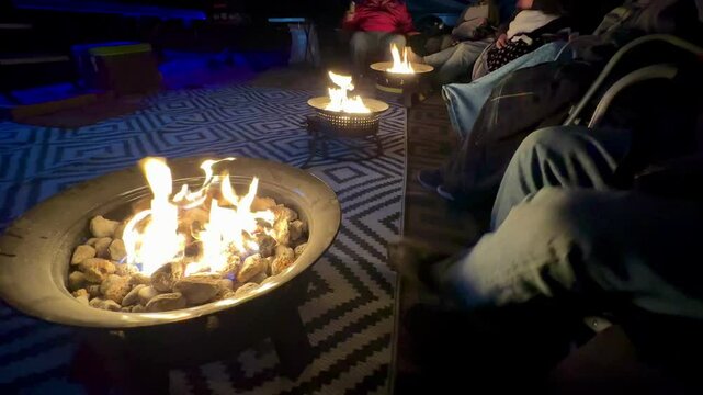 Cozy campsite gathering features diverse group of outdoor enthusiasts relaxing around portable propane fire pits, enjoying warmth and camaraderie amidst a natural setting.