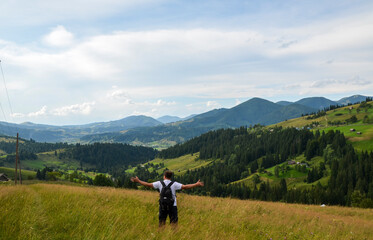 Obraz premium Serene landscape featuring a man with arms outstretched in a lush mountain valley, surrounded by vibrant green forests and rolling hills under a picturesque clear sky. Carpathians, Ukraine