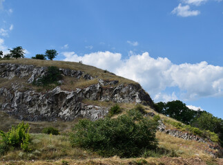 Blue sky with clouds above hill with trees, grass, bushes and rocks.
