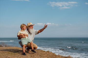Grandfather teaching his grandson about the world, pointing at the ocean horizon on a sunny day,...