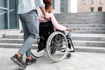 Obraz premium Young black guy pushing wheelchair with handicapped woman, having problem entering building, standing in front of stairs without ramp. Problems with accessible environment for disabled people