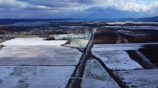 Panoramic drone view of Sayabec village and its straight railway line beside Lake Matapedia under cloudy skies in the snowy agricultural landscape of the Matapedia Valley, Quebec, Canada