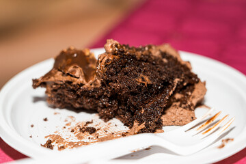 Close-up of a slice of chocolate cake with multiple layers, served on a white plastic plate alongside a plastic fork. Captured on a vibrant pink tablecloth.