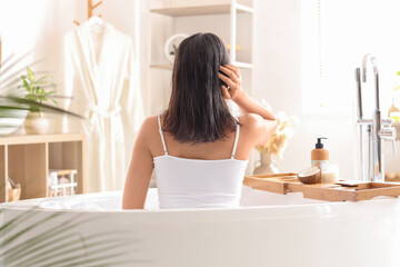 Pretty young woman applying coconut oil on her hair in bathroom, back view