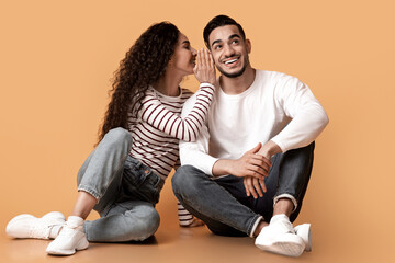 Sharing Secrets. Cheerful Young Woman Whispering To Her Boyfriend's Ear While They Sitting On Floor Over Yellow Background In Studio, Happy Middle Eastern Couple Gossiping Together, Copy Space