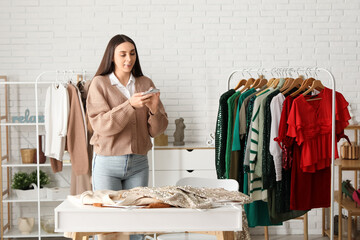 Happy female stylist with clothes on rack taking photo of dress at table in studio