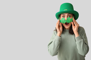 Shocked young woman in leprechaun hat and decorative glasses in shape of clover on grey background....
