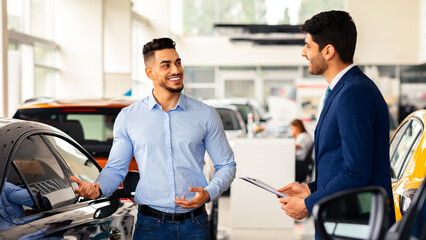 Cheerful arab guy standing by nice black sports car, talking to dealer, side view. Wealthy middle-eastern young man having conversation with handsome sales manager at luxury showroom