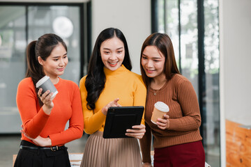 Three young women in a modern office, smiling and collaborating over a tablet, holding coffee cups, showcasing teamwork and technology.