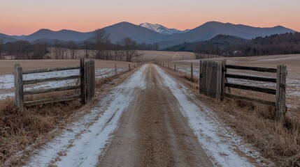 Fototapeta premium Dirt road with patches of snow leads through wooden gates toward distant mountains under a pink twilight sky.