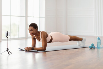 Smiling African American lady in white sportswear doing plank, shooting fitness video or having online live chat with coach, sporty lady exercising and looking at mobile phone screen on tripod