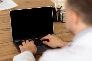 Lifestyle And Gadgets Concept. Over the shouder view of adult man using laptop with blank black screen for mockup template, sitting at wooden table, back shot. Person typing on computer keyboard