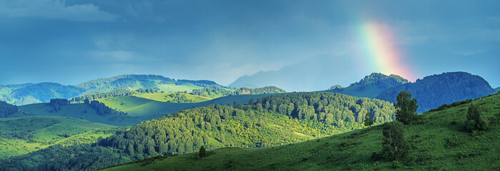 View of green meadows and hills on a summer evening, stormy sky and rainbow, panoramic view