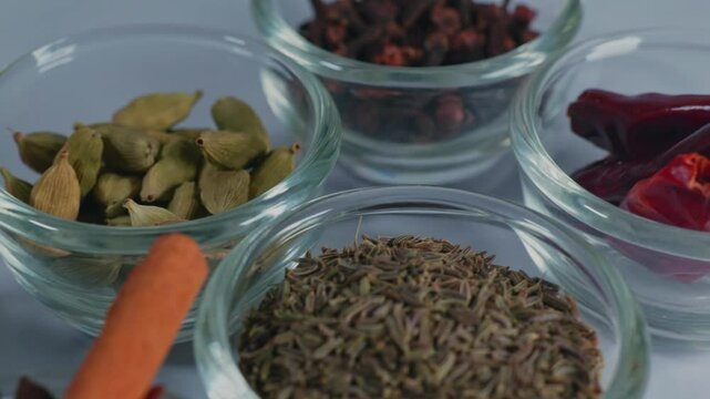 different types of whole spices Cinnamon stick, Black pepper, Caraway seed, green cardamom, dried red chilis and Cloves in small glass bowls, 4k