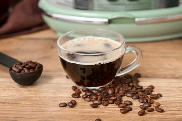 Glass cup of hot espresso with beans and modern coffee machine on wooden background, closeup