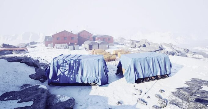 A remote research station is situated in a snowy landscape, with multiple buildings shrouded in white. Covered equipment rests among rocks, highlighting the isolation and wintry environment.