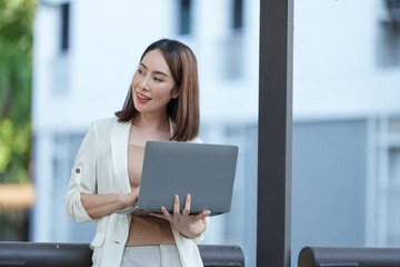 Asian businesswoman standing outdoors, using a laptop computer while smiling and looking away, embodies confidence and professionalism in a modern urban environment