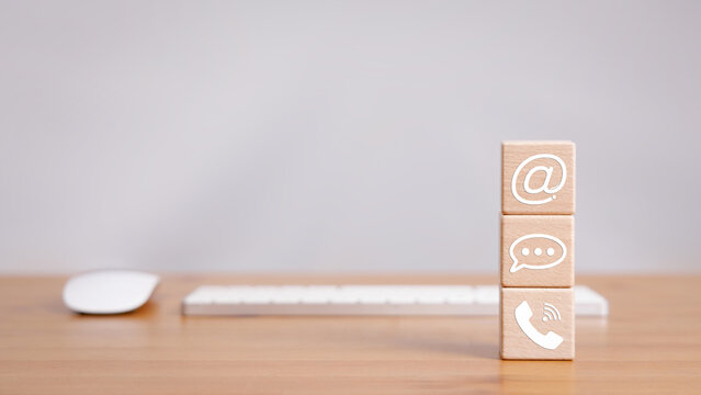 Cube wood block stacked on wooden table with phone, message bubble, and email sign icon with computer keyboard and mouse in contact us concept.