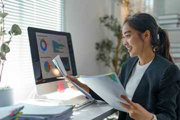 Young office worker smiling while analyzing financial charts and graphs, holding documents and...