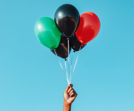 an African American hand holding a bunch of green, black, and red balloons against a clear blue sky. Black Liberation African American Flag. Pan-African flag. Juneteenth National Independence Day. 