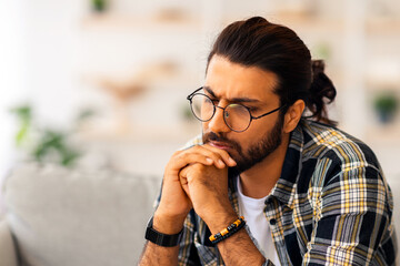 Pensive long-haired indian guy with glasses sitting on couch at home, looking for solutions,...