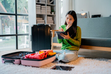 Asian woman writing notes in notebook while preparing clothes for travel, packing suitcase on floor at home