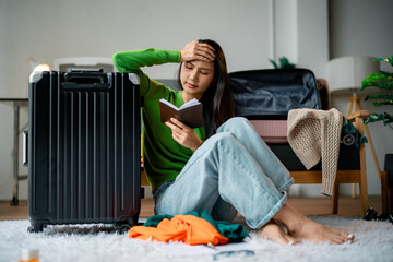 Stressed young woman sitting on the floor at home, packing a suitcase and holding a passport,...