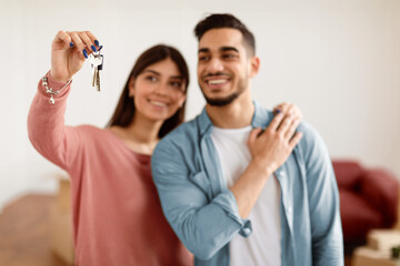 Positive young couple showing house key, standing in living room of new home, selective focus hand....