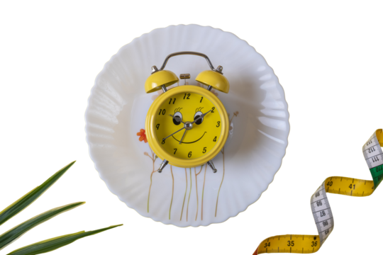 a white plate with a floral pattern, on which stands a yellow alarm clock with a smiling face. The alarm clock has two bells on top and arrows pointing to 10:10. There are green leaves in the lower le